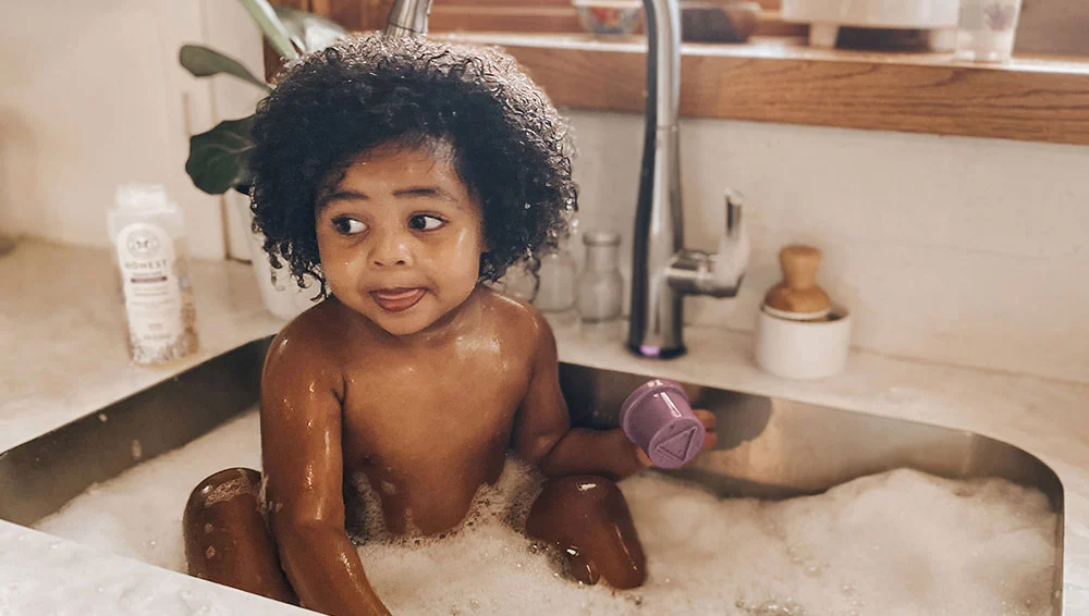 African american baby taking a bath in a kitchen sink using The Honest Compnay products